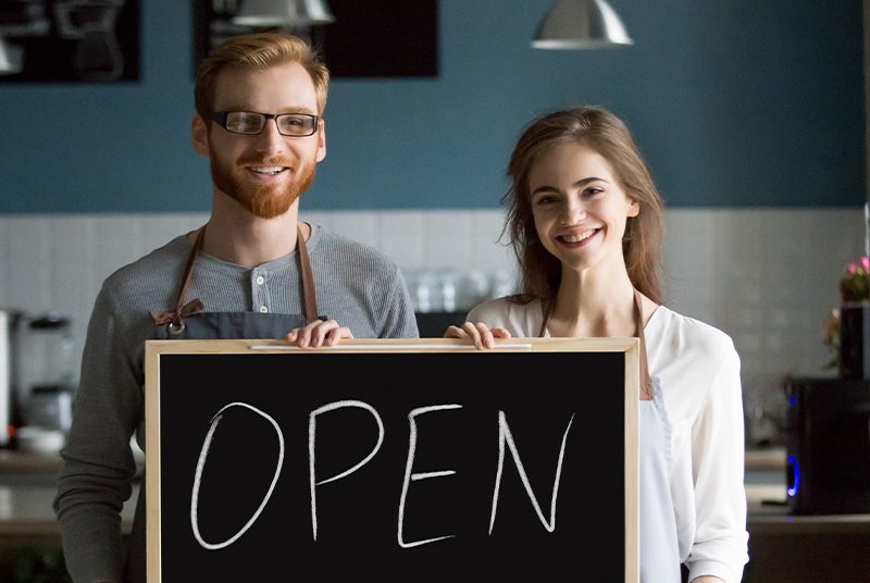 A man and woman in a cafe hold an 'open' sign|A man and a woman serving drinks at a bar|A chef working in a kitchen dealing with food orders
